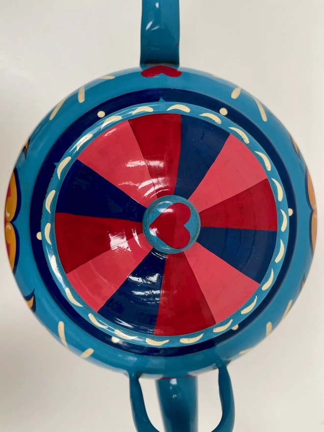 The lid of a large blue enamel teapot hand painted with a traditional fairground design. The lid is painted with blue, red and pink stripes.