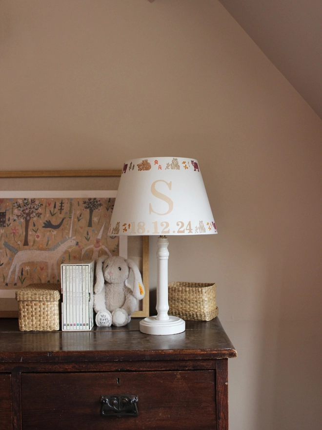 Cozy corner of a girls bedroom, featuring a wooden dresser and white table lamp paired with a personalised kids lampshade adorned with a bears pattern and the letter 'S'. A framed artwork hangs behind the lampshade featuring a unicorn and mythical setting.