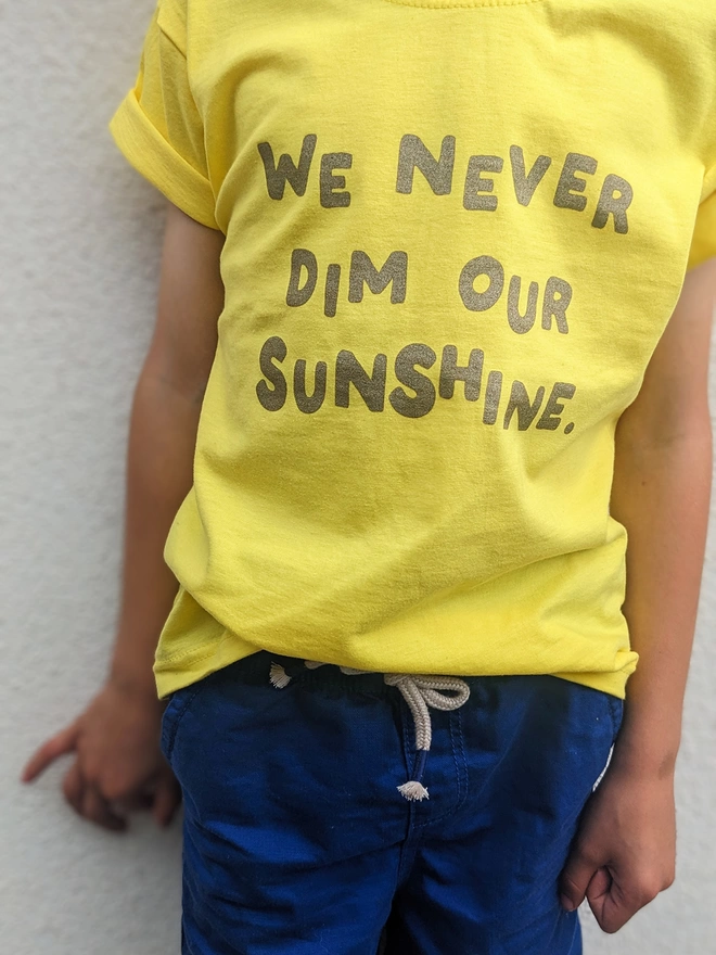 A child by a white wall wearing a yellow t-shirt with We Never Dim Our Sunshine t-shirt