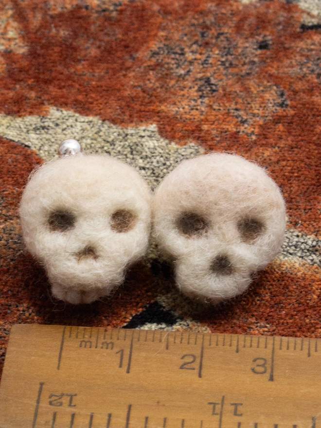 Two fuzzy needle-felted skull earrings on an orange autumnal fabric. Made by the artist Laura Cronin aka Bumble & Earwig.