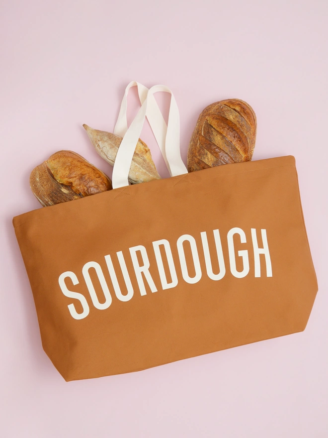 oversized tan canvas tote bag with sourdough slogan laying on a pink background with bread spilling out model carrying an oversized tan canvas tote bag with sourdough slogan