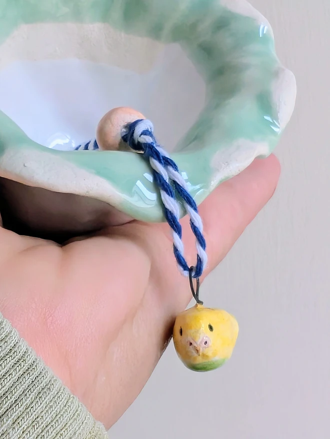 white budgie bell in ceramic with a tiny yellow and green budgie held in a hand to show a close up of the bird