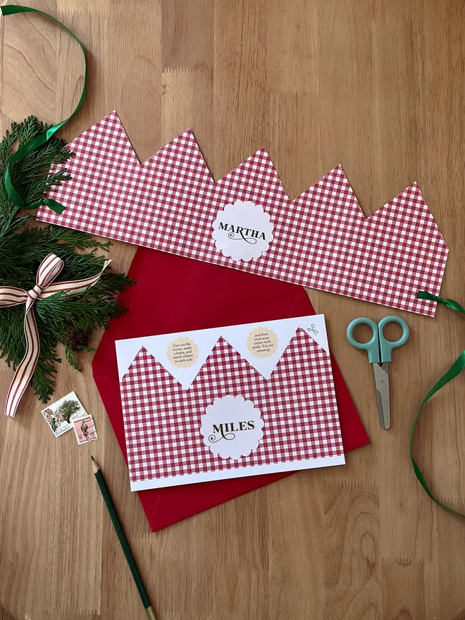 A Christmas crown card with a red gingham pattern lays on a red envelope, on a wooden table.