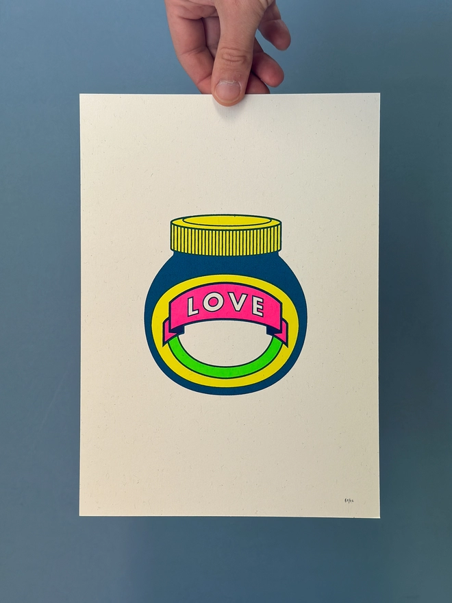 Hand holding an A4 screen print showing the icon design of the famous Marmite jar with the word LOVE reworked into the jar label. The design is made up of 4 flouro colours. 