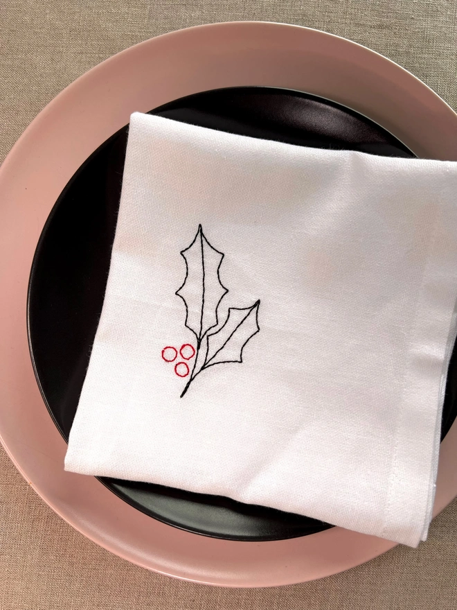 White cotton embroidered Christmas holly napkin, styled on pink and black place setting
