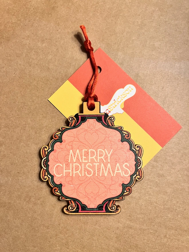 Multi coloured wooden tree decoration with 'Merry Christmas' written at the centre. It has a red ribbon and is lying on a brown card background with a business card with Rebecca Strickson on it. 