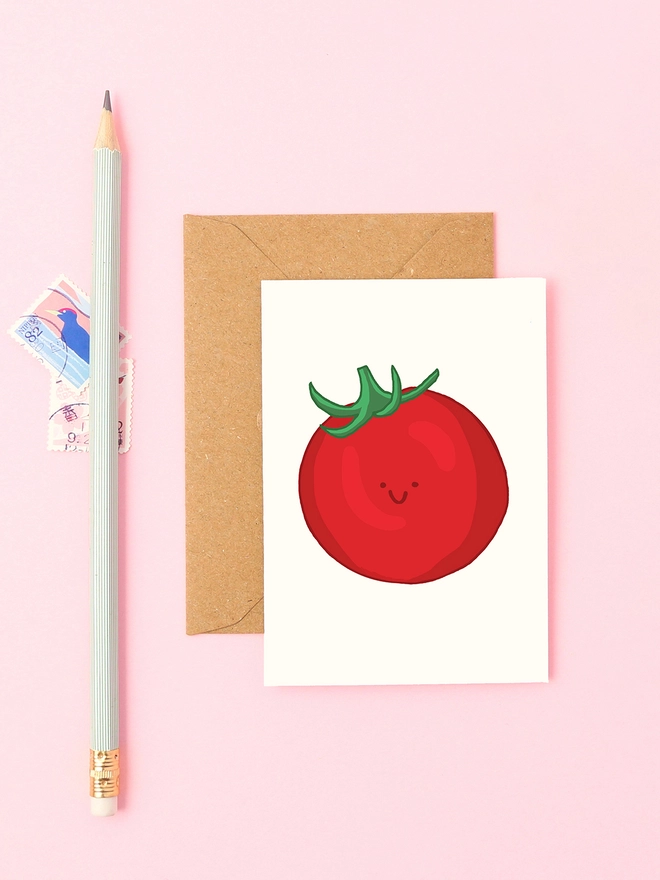 Mini greeting card with a hand-drawn smiling tomato illustration, styled with a kraft envelope, stamps and a mint pencil on a pastel pink background