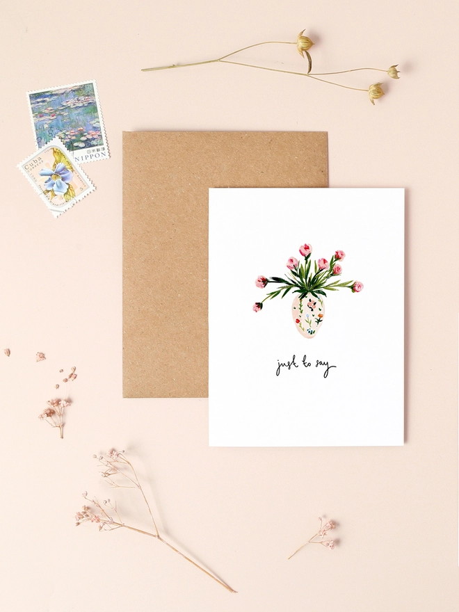 An image of a mini, rectangular white greetings card with an illustration of a vase of pink flowers with a hand written 'just to say' message in black ink seen under the central illustration. The card is placed on top of a brown rectangular kraft envelope.