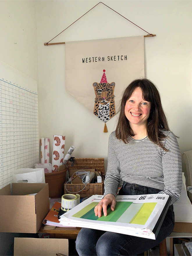 Illustrator Melissa Western Sitting On Her Studio Desk Holding Her Sketch Book.  Behind Her Is Her Western Sketch Business Banner Which Features A Hand Appliqué and Embroidery Of Her Leopard Illustration.