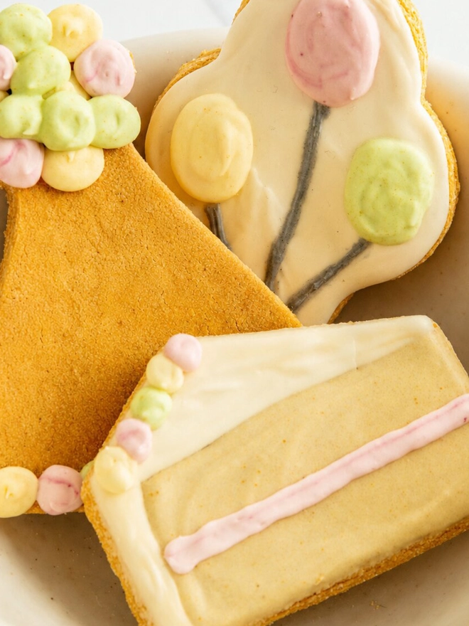 Close-up of cake slice, party hat and balloon bone Celebration iced dog biscuits arranged in a ceramic bowl.
