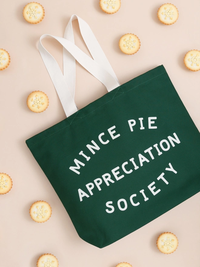 Green canvas Mince Pie Appreciation Society tote bag laying on a coloured backdrop surrounded by mince pies