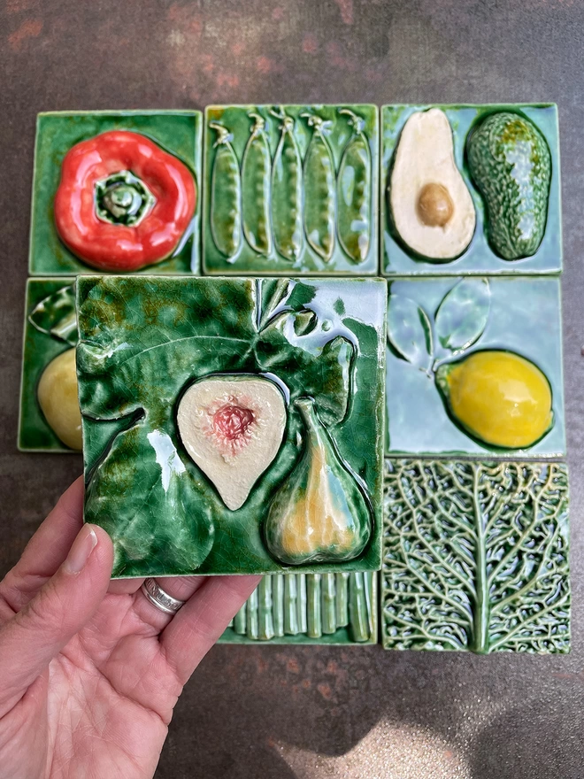Fig tile - square, 3D, realistic and glossy. The fig is cut open to reveal a fleshy pink inside, and the leaf has a lush green glaze. Other fruit and vegetable tiles in the series are on display in the background: lemon, savoy cabbage, red capsicum pepper, pear, mange tout, avocado, garlic, asparagus.