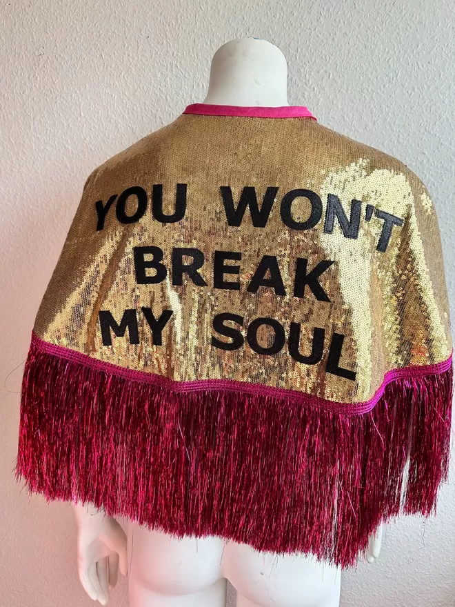 a maxi cape with gold sequins, gold text reading 'YOU WON'T BREAK MY SOUL' and hot pink tinsel