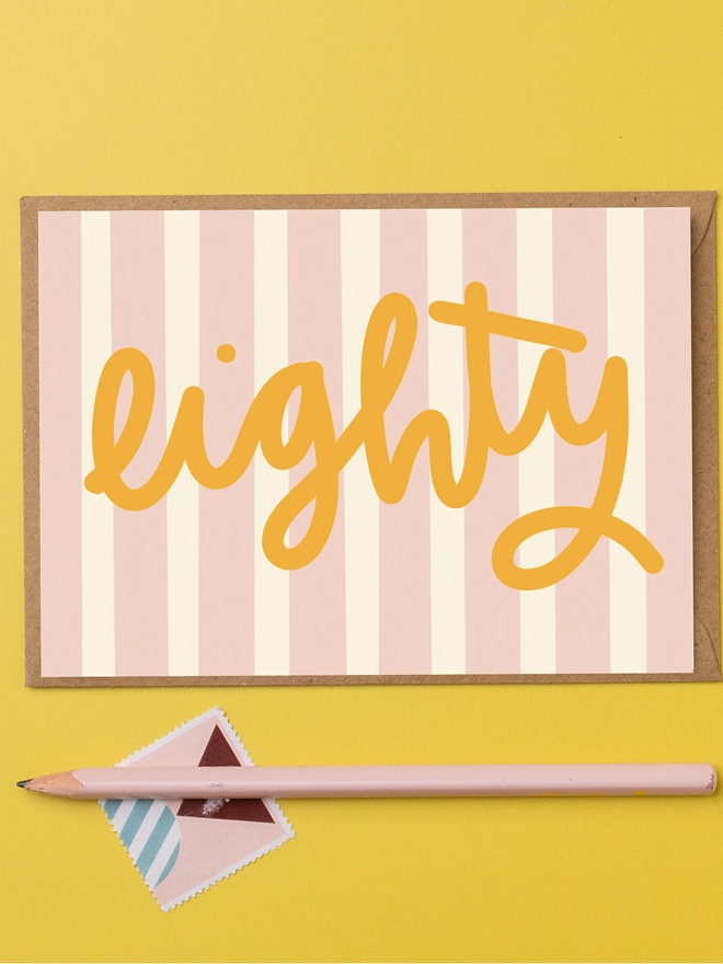 An 80th birthday card featuring the word eight in mustard yellow lettering on a pink and cream striped background