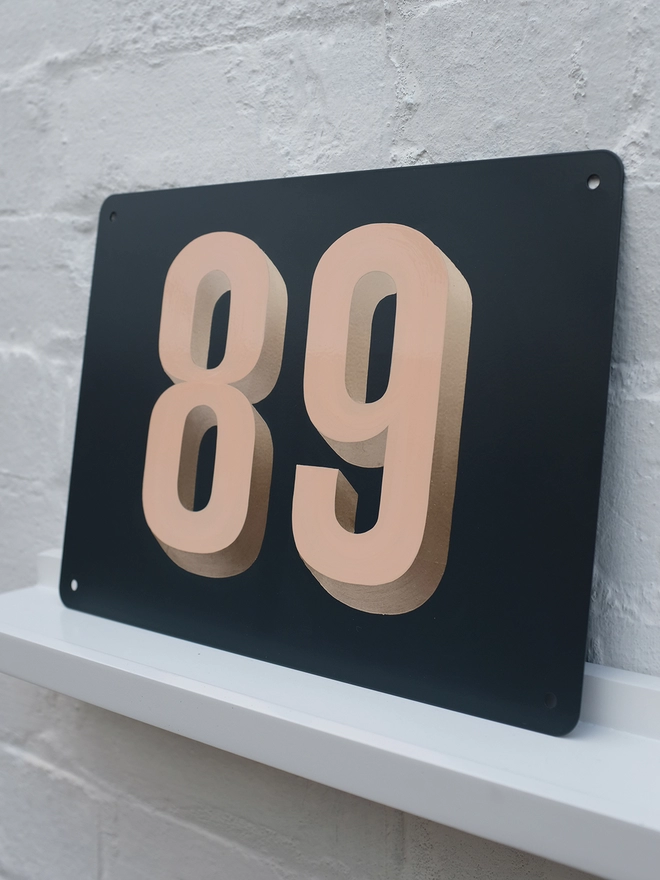 Peach and gold leaf house number 89, on anthracite grey metal plaque, against a white brick wall.