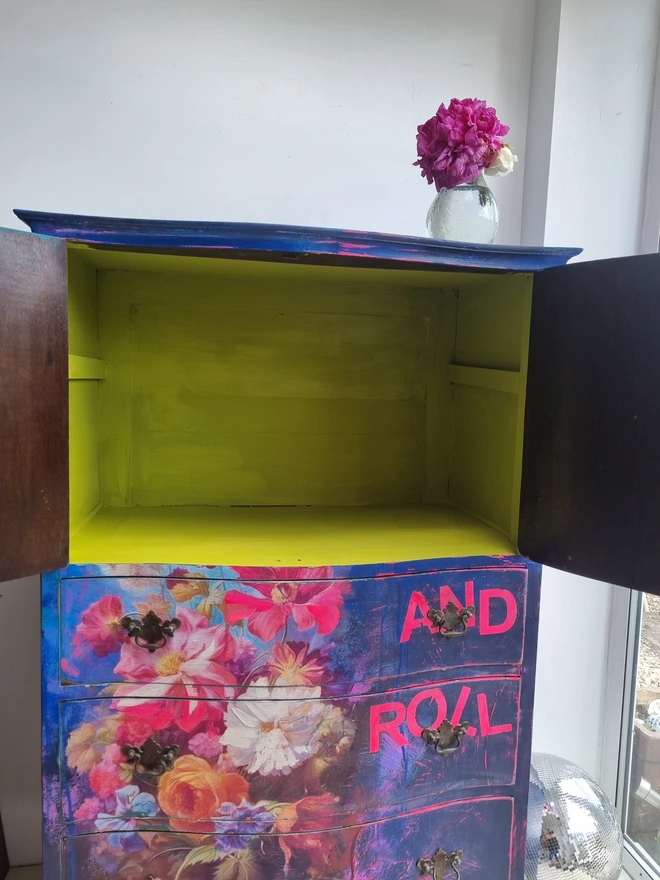 A vintage tallboy cabinet painted in vibrant blues, neons and flowers with the text that reads 'we built this city on rock and roll' in neon pink.