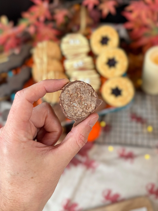 A hand holds a round chocolate-covered welshcake ball in the foreground, with more decorated welshcakes blurred in the background on a Autumn styled tabletop.