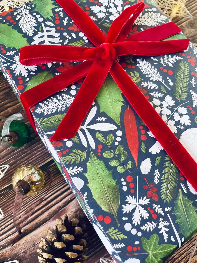 Close-up of present wrapped in Winter leaf wrapping paper with Holly and Ivy design, red bow, surrounded by star lights, baubles, and pine cone on wood background