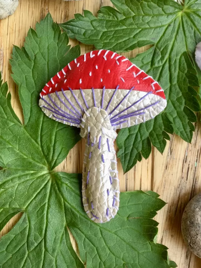 Three hand stitcheA red and white hand stitched faux leather toadstool brooch laying amongst leaves on a wooden surface. 