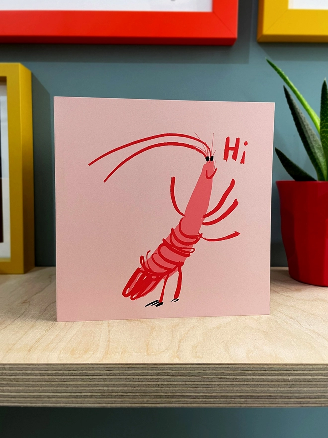 A happy cartoon prawn says Hi on this jolly upbeat pink card, printed in two pink inks with black eyes. Stood on a plywood shelf and other framed pictures showing around the edges. A plant is half in shot.
