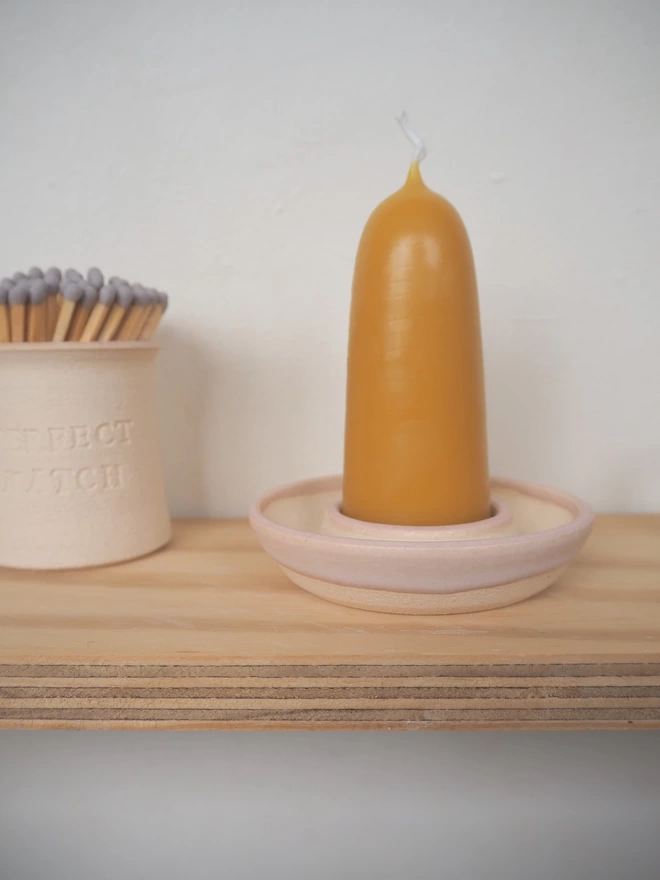 Wooden shelf with pale pink candle holder and short stubby beeswax candle. Nect to it a match pot containing grey tip matches