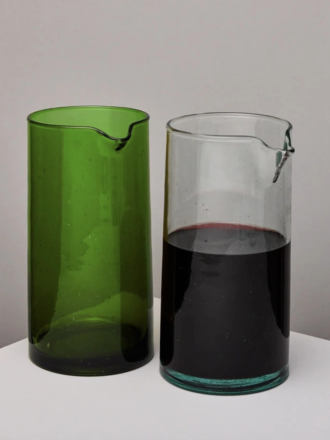 Glass Carafe, a large green glass jug sitting on a plain counter top against a grey backdrop. It is accompanied by a clear carafe filled with wine. 