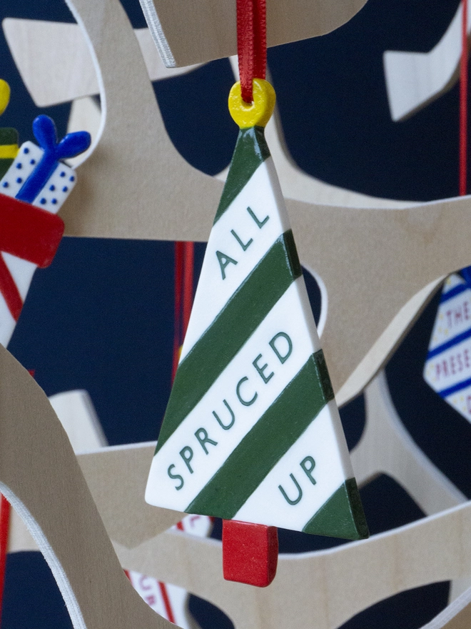Ceramic porcelain Christmas decoration with All Spruced Up written on the side. A white tree-shaped Christmas decoration with green lettering, decorated with green stripes and hanging from a plywood Christmas tree.