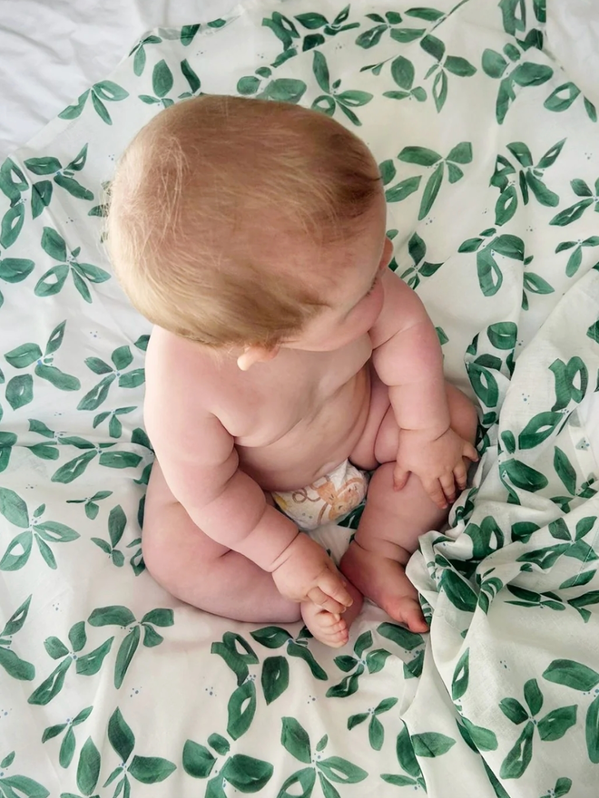 Emerald Leaves Muslin. A muslin patterned with green leaves, with a baby sat on top.