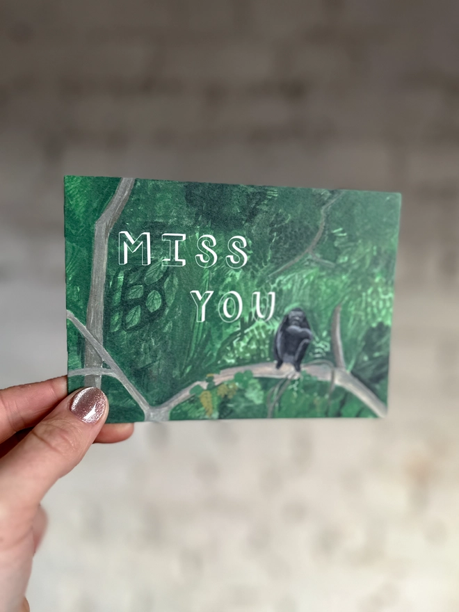 a greetings card featuring an illustration of a chimp sitting in a jungle landscape with the text “miss you”