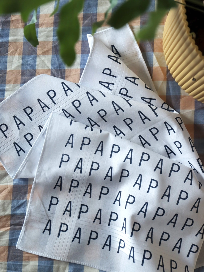 A Mr.PS PAPA hankie on a gingahm tablecloth with a yellow plant pot and leaves