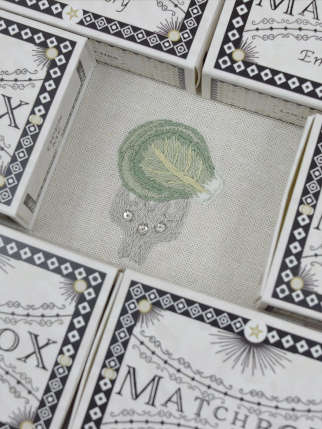 an embroidered Christmas Brussels Sprout on a fork studded with crystals, surrounded by matchbox embroidery kits