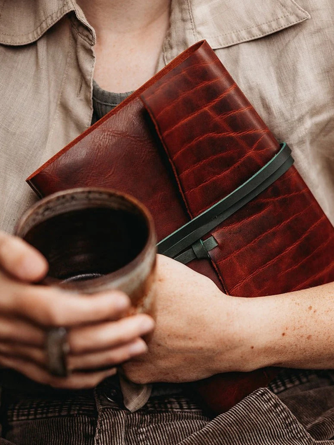 make your own leather journal. a red-brown leather journal held in one hand with a cup of coffee in the other.