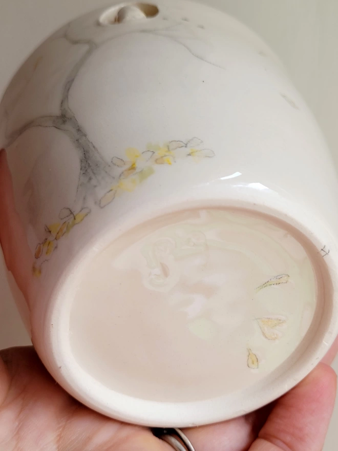 ceramic white cup held in a hand to show the base with painted leaves and heart there is a painted tree and leaves on the ground on the beaker