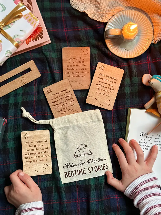 A personalised cotton bag of wooden bedtime story prompt cards is displayed on a cosy red tartan blanket, featuring engraved story ideas. A child's hands are seen holding the cards.