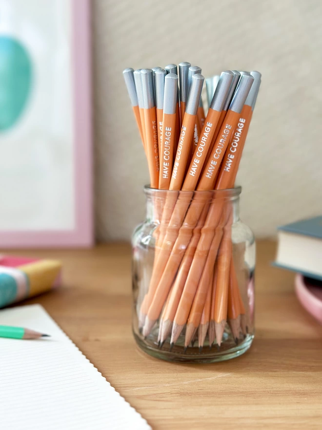 A glass jar full of orange pencils stands on a wooden desk surrounded by stationery items. Each pencils has the words Have Courage on the side.