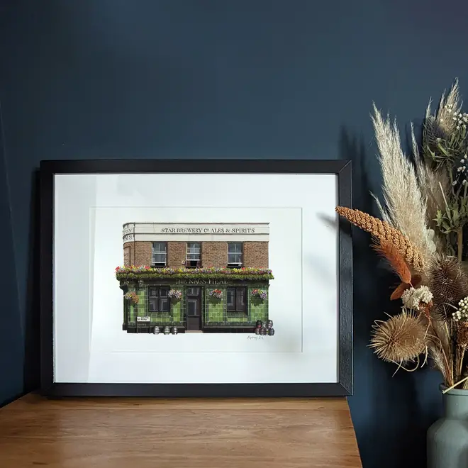 Greenwich The Pelton Arms Pub Giclée Print (Unframed), a print of a brown and green pub sitting against a navy blue backdrop. It is accompanied by some flowers
