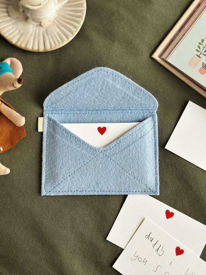 A handmade envelope lays open on dark green fabric. Tucked inside are mini heart notecards, with several written cards beside it. Each written card has a note to Daddy written by children.