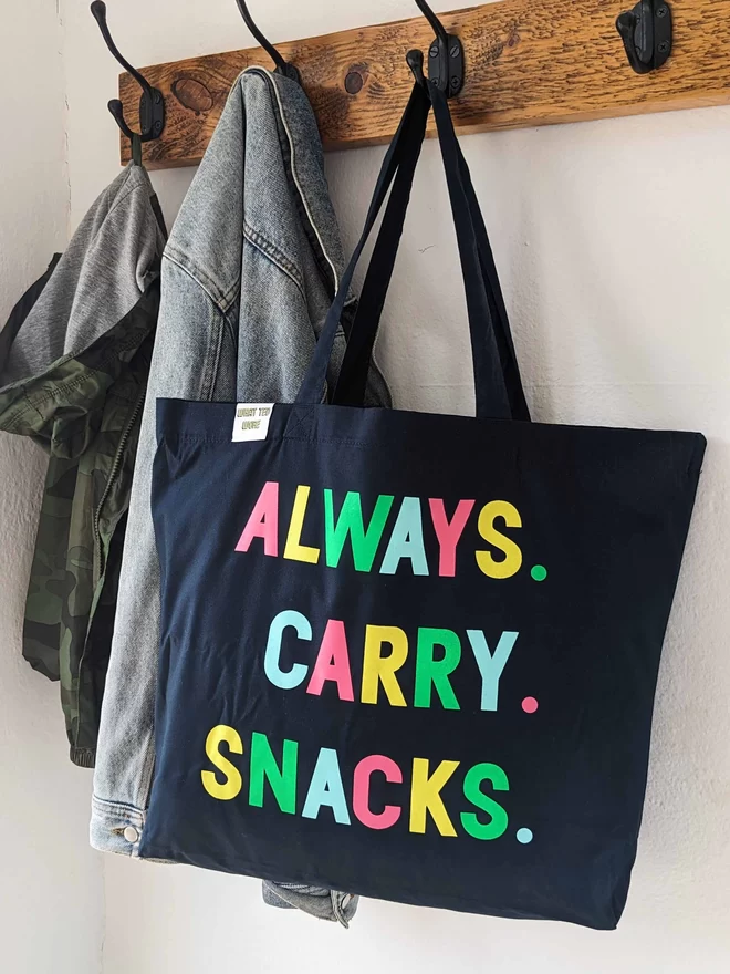 A hanging navy canvas tote bag with Always Carry Snacks slogan