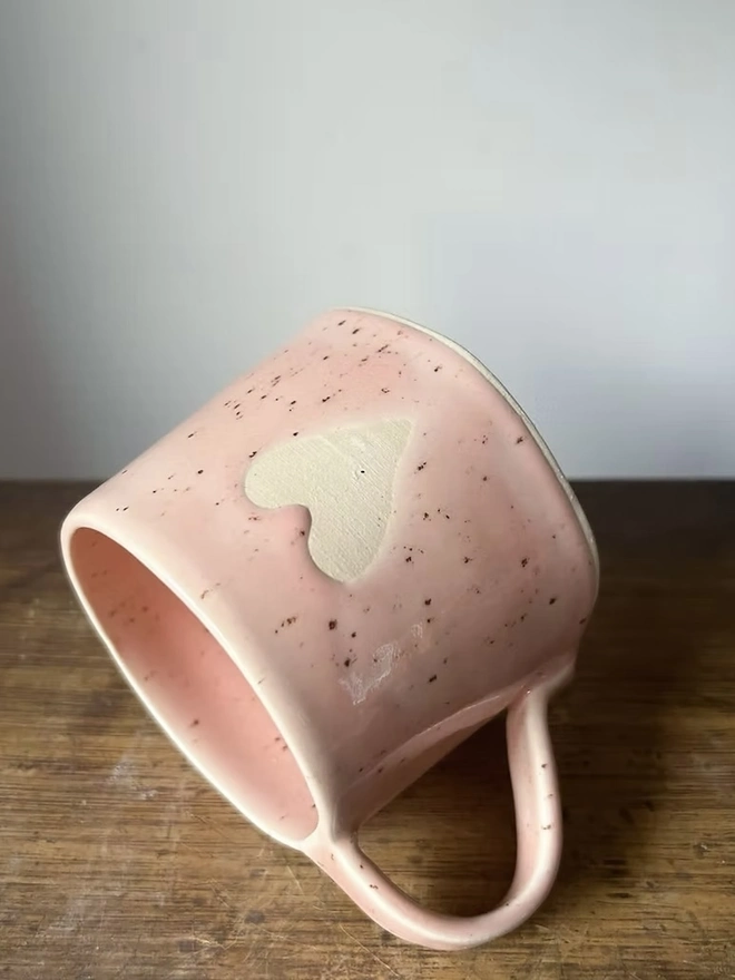 Heart Mug Strawberry Pink, on a wooden surface lying on its side in front of a white background. 