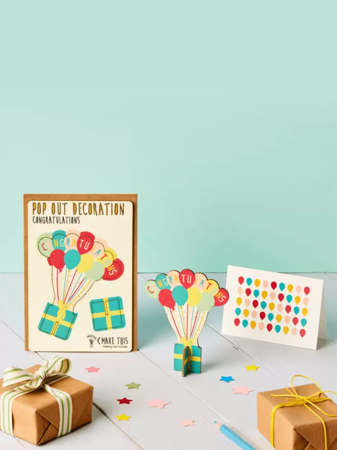 3D congratulations decoration and balloon pattern greeting card and brown kraft envelope on top of a grey desk in front of a light green coloured background
