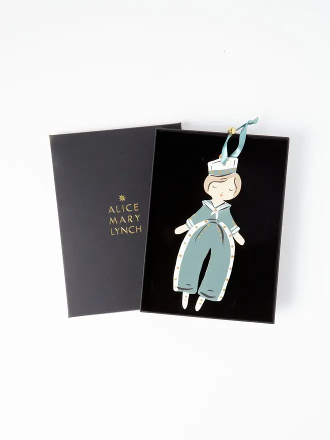 Sailor Heart Tree Dancer. wooden hanging sailor boy decoration in an open black gift box embossed "alice mary lynch" in gold, with a white sailor hat, sage green and white jumpsuit with gold buttons