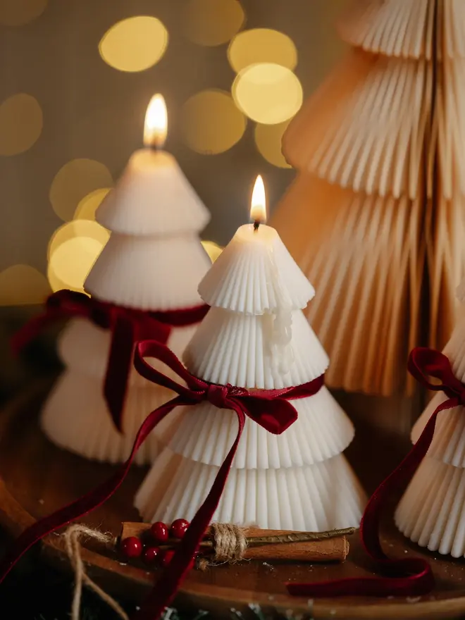 Tiered Christmas Tree Candle Close-up of a glowing cream Christmas tree candle with fine ribbed layers and a deep red velvet bow, beside cinnamon sticks and berries. The detailed sculptural design adds a soft, romantic glow to winter evenings.