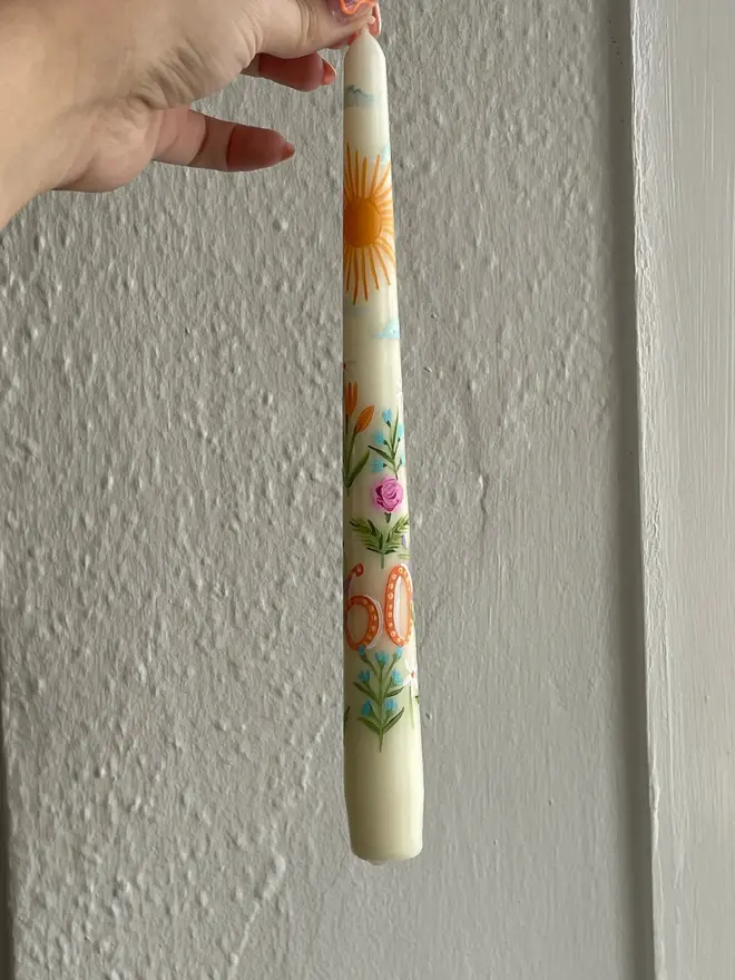 Birthday Or Anniversary Hand Painted Candle, a person holding the birthday or anniversary candle against a white surface. The '60' can be seen next to the floral design wrapping around it. 