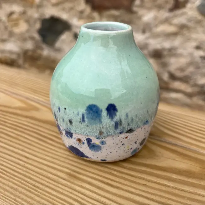 Shoreline Bud Vase, a colourful vase placed on a wooden surface against a brick backdrop. 