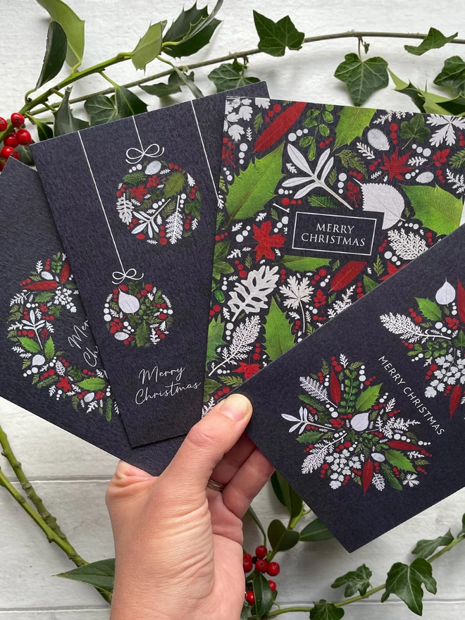 Hand Fanning Out 4 Christmas Cards with Red, White, and Green Pressed Winter Leaf Designs - Sprigs of Holly on White Background
