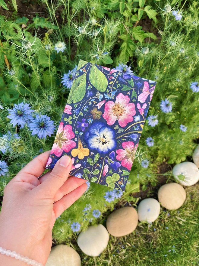 Hand Holding Pretty Notebook with Pressed Flower Design, Blue Nigella Flowers, Foliage, and Pebbles in Garden Background