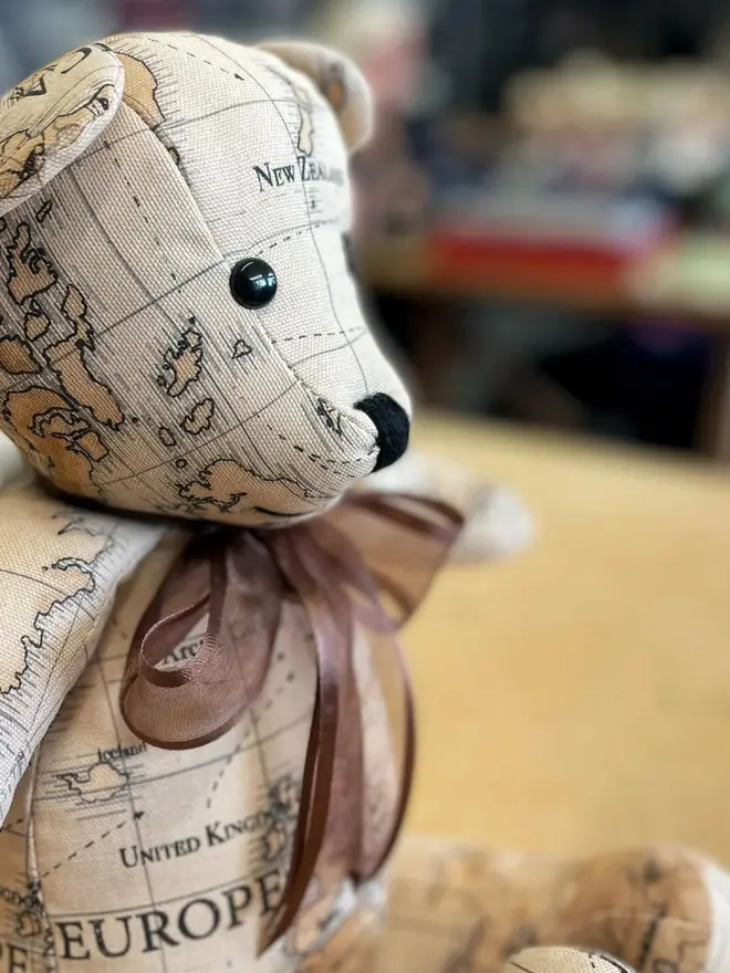 Teddy bear made from map-print fabric sitting upright on a wooden table, wearing a brown bow.