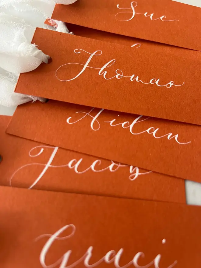 Personalised Calligraphy Rust Orange Place Card, orange place cards with white chiffon ribbons grouped together on a white surface. 