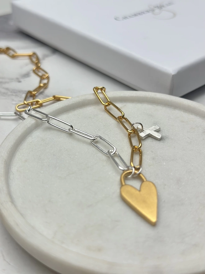 an elongated gold heart charm hangs from a mini paperclip chain which is half silver and half gold.  Hanging 3 links up the chain is a small silver x shaped kiss charm