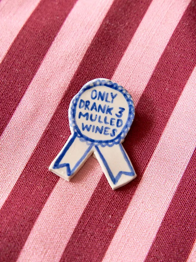 'Only Drank 3 Mulled Wines' hand-painted on a ceramic Christmas award badge in blue, atop a striped burgundy and light pink tablecloth